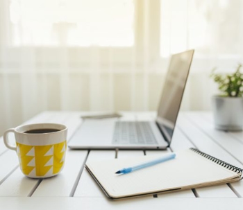 A work-from-home set up next to an open window with sunlit curtains featuring an open laptop next to a notebook with a pen and a mug of coffee.